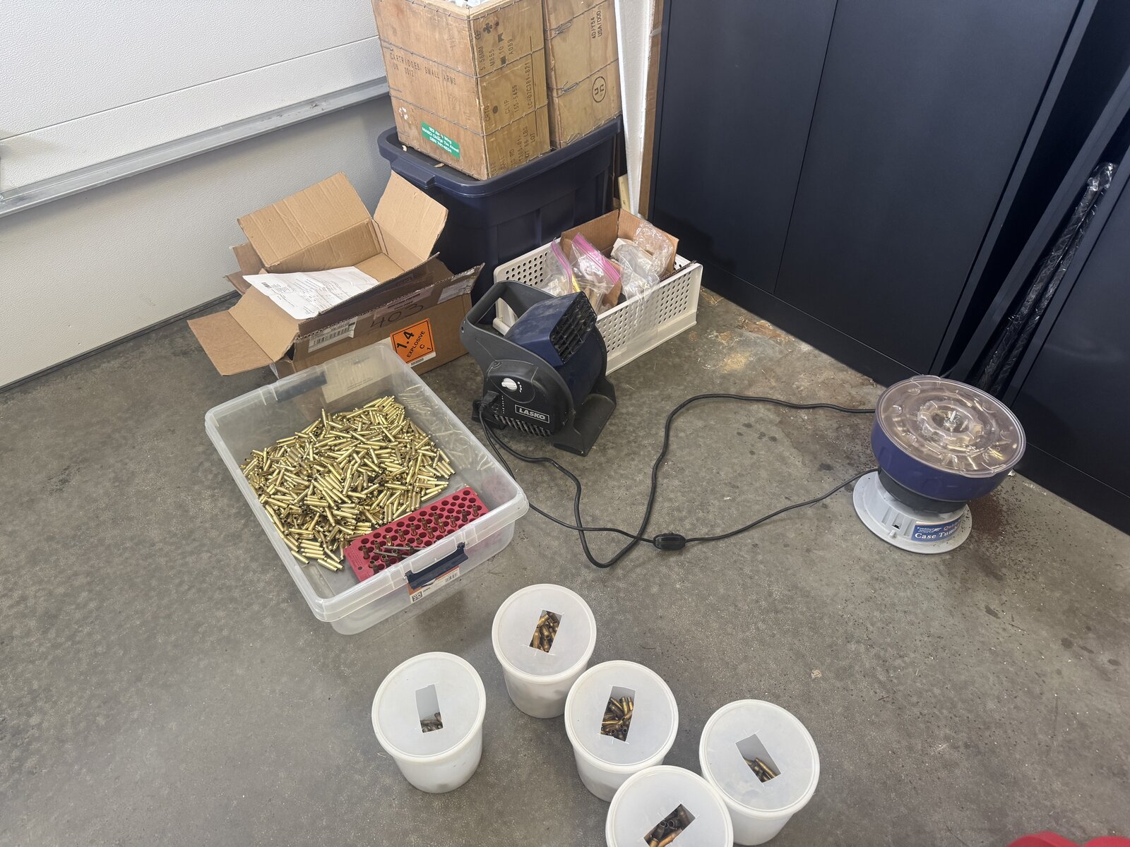 Labeled buckets, a tub of mixed brass, ammunition crates, and a case tumbler arranged on the garage floor