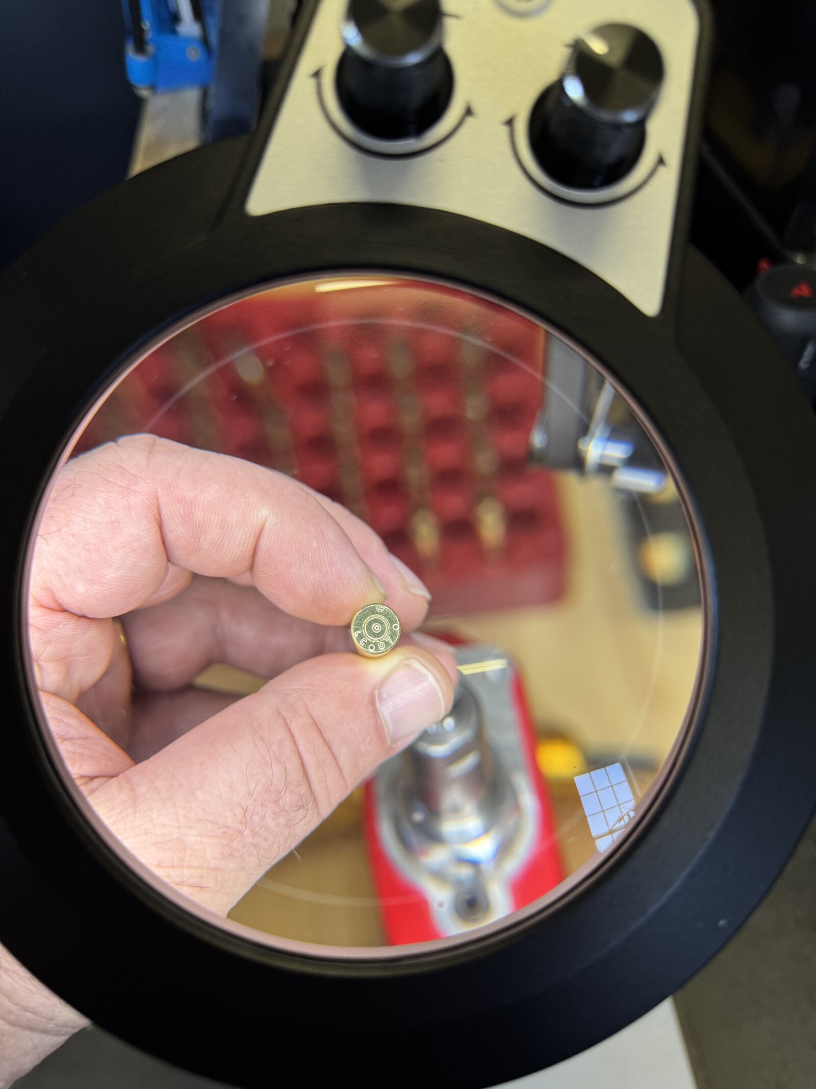 Fingers holding a cartridge case head under a lighted bench magnifier, the headstamp legible through the lens